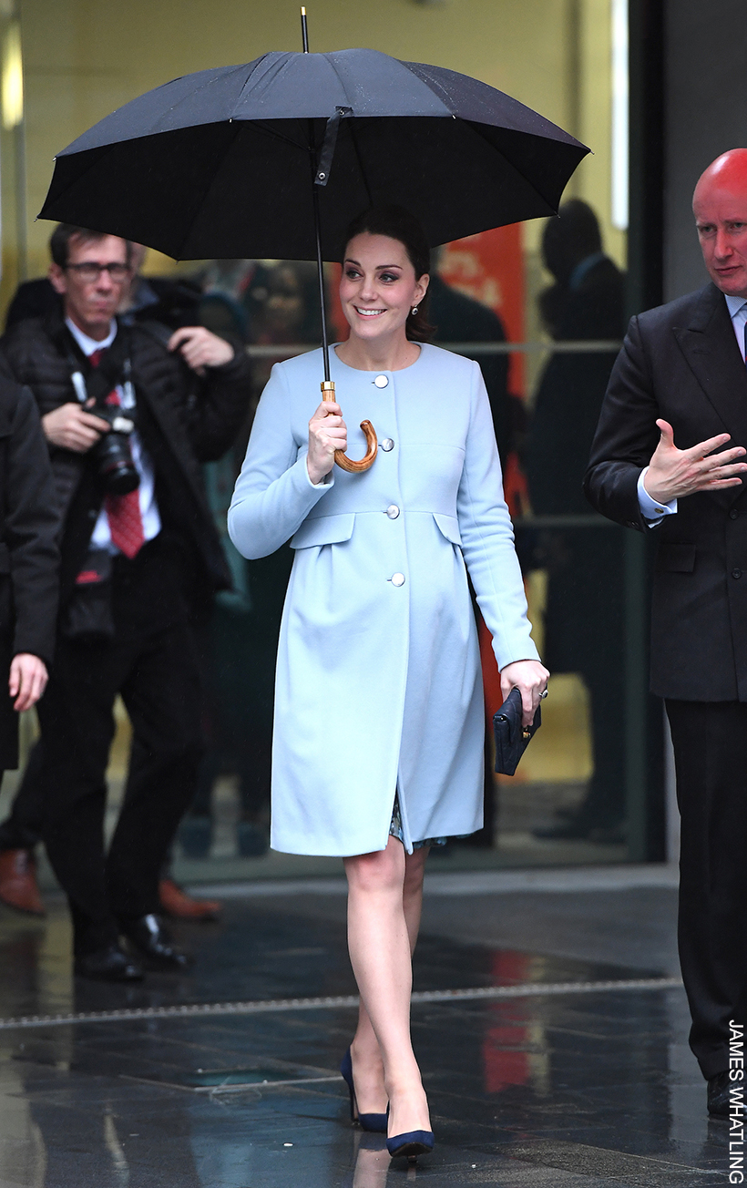 Kate Middleton walks out of a building holding an umbrella wearing a knee-length blue maternity coat.