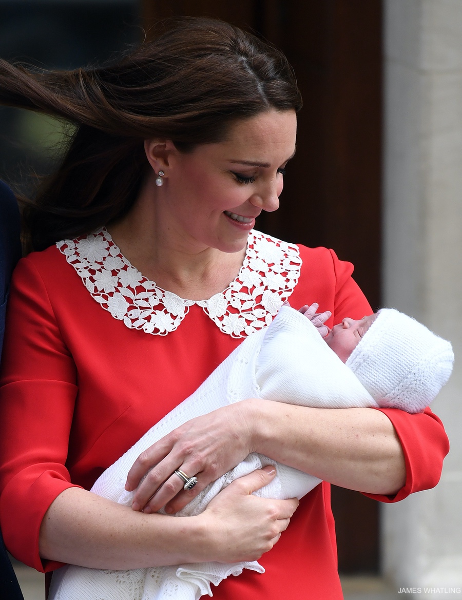 Kate Middleton presents her new baby boy to the world! The Princess, grinning, wears a red dress with a white lace floral collar, designed by Jenny Packham, 