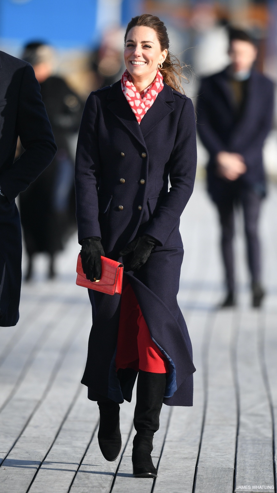 Kate Middleton wearing the ankle-length long blue wool maxi coat from Hobbs London.  The coat is double-breasted with metal-tone buttons.  The Princess wears the coat with red accessories. 