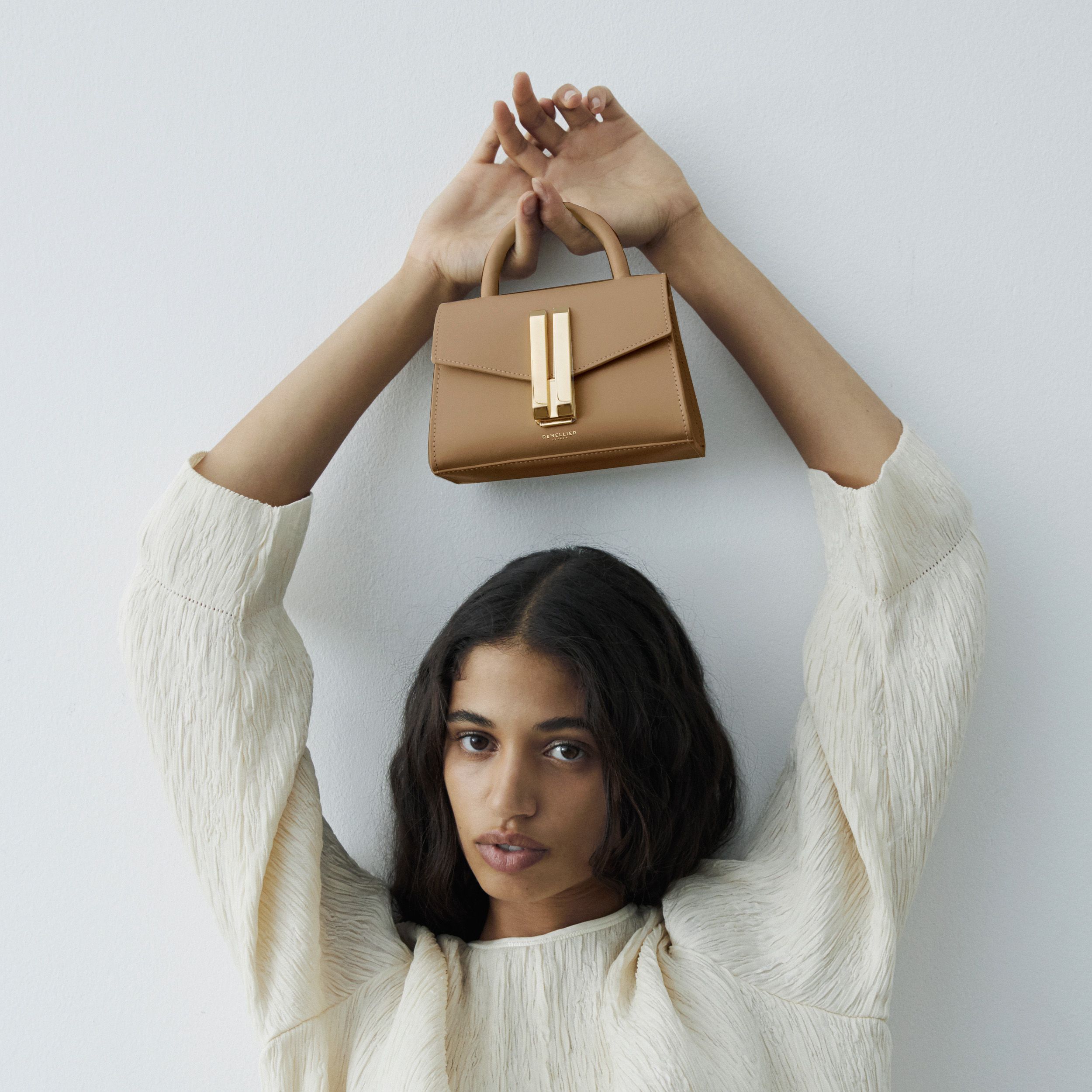 Model posing, holding the toffee Demellier handbag above her head.  Model is wearing a white top. 