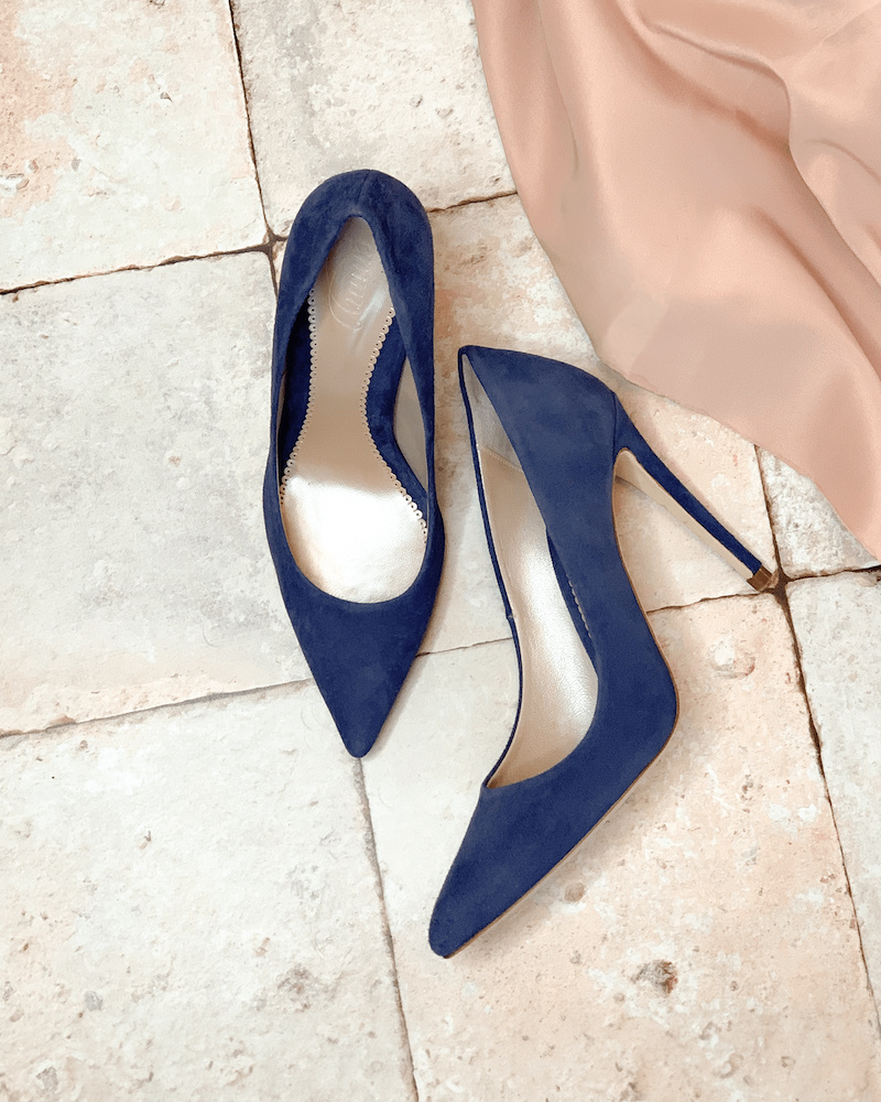 A pair of Emmy London Rebecca Court Shoes in Riviera Blue Suede placed on a neutral-toned tiled floor, with a blush pink fabric draped in the background.