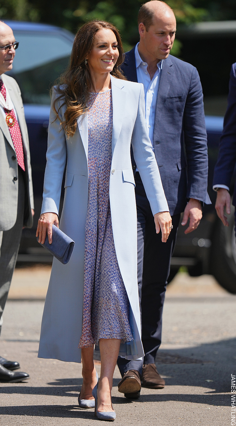 Kate Middleton walking outdoors, wearing the Emmy London Rebecca Court Shoes in Riviera Blue Suede, paired with a light blue coat, lilac printed dress, and a matching clutch bag.

