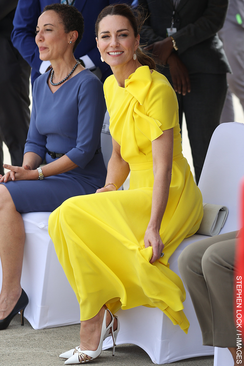 The Princess of Wales sitting, smiling, wearing a yellow dress with a large bow on the shoulder and mother-of-pearl earrings.  Her striking white and gold shoes are on display.