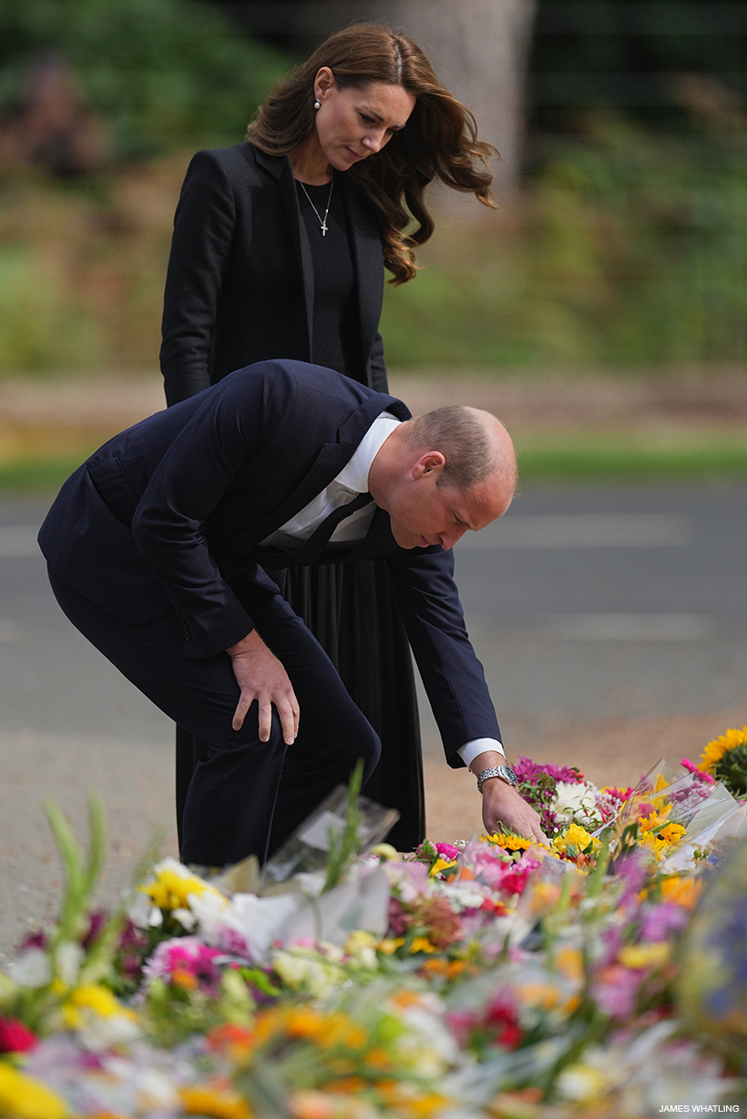 Prince William bends down to read a card left beside a floral tribute to his late grandmother.  Kate stands behind him.