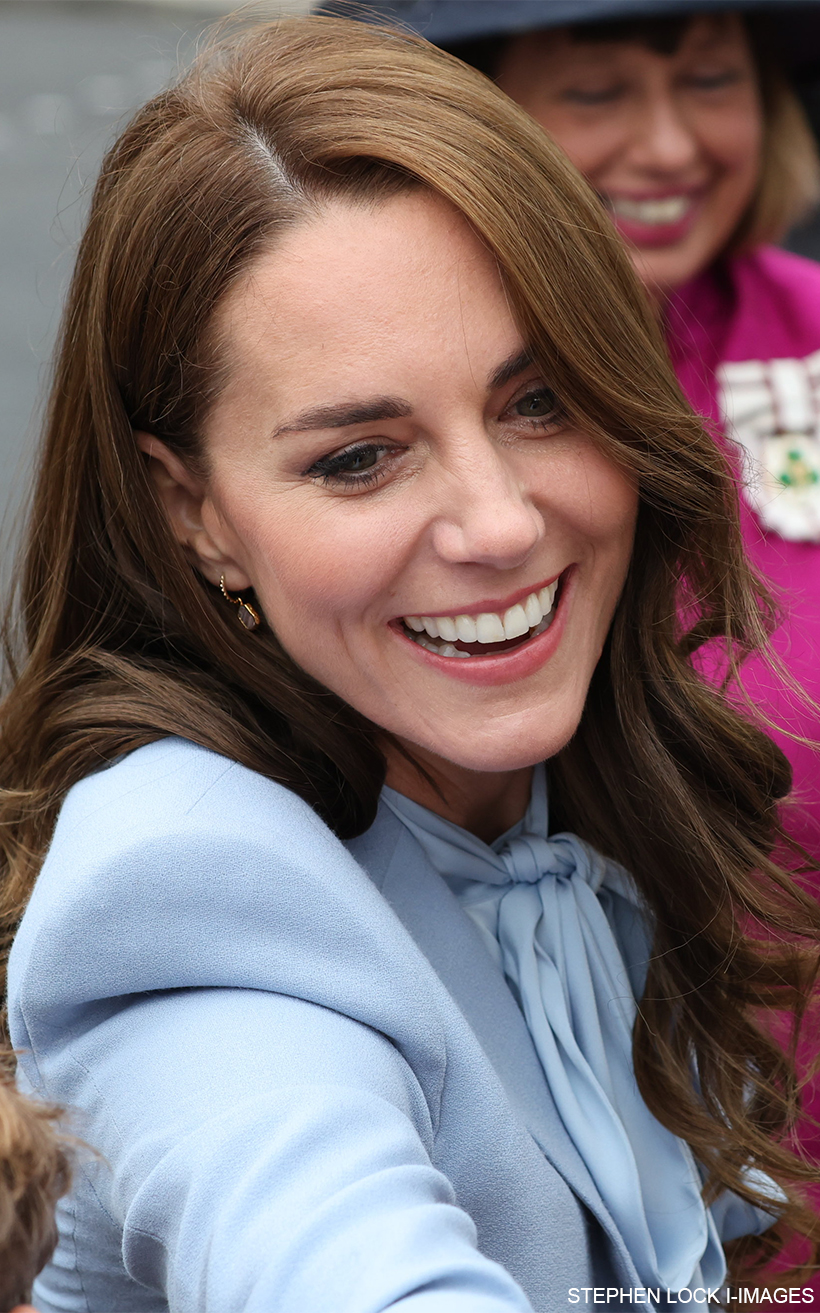 Close up of Kate Middleton smiling and greeting a member of the public in the crowd.  She's wearing her blue coat, blue bow blouse and pink/gold earrings.