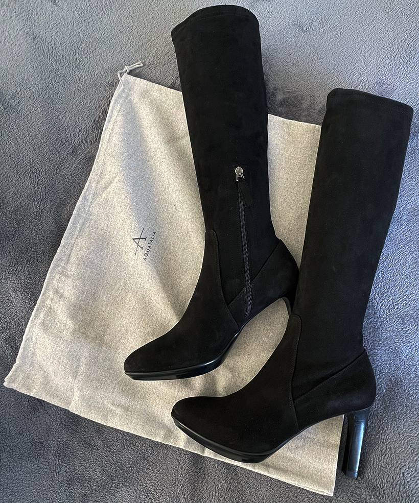 Black suede Aquatalia Rhumba boots lying on their dust bag, on a grey background.