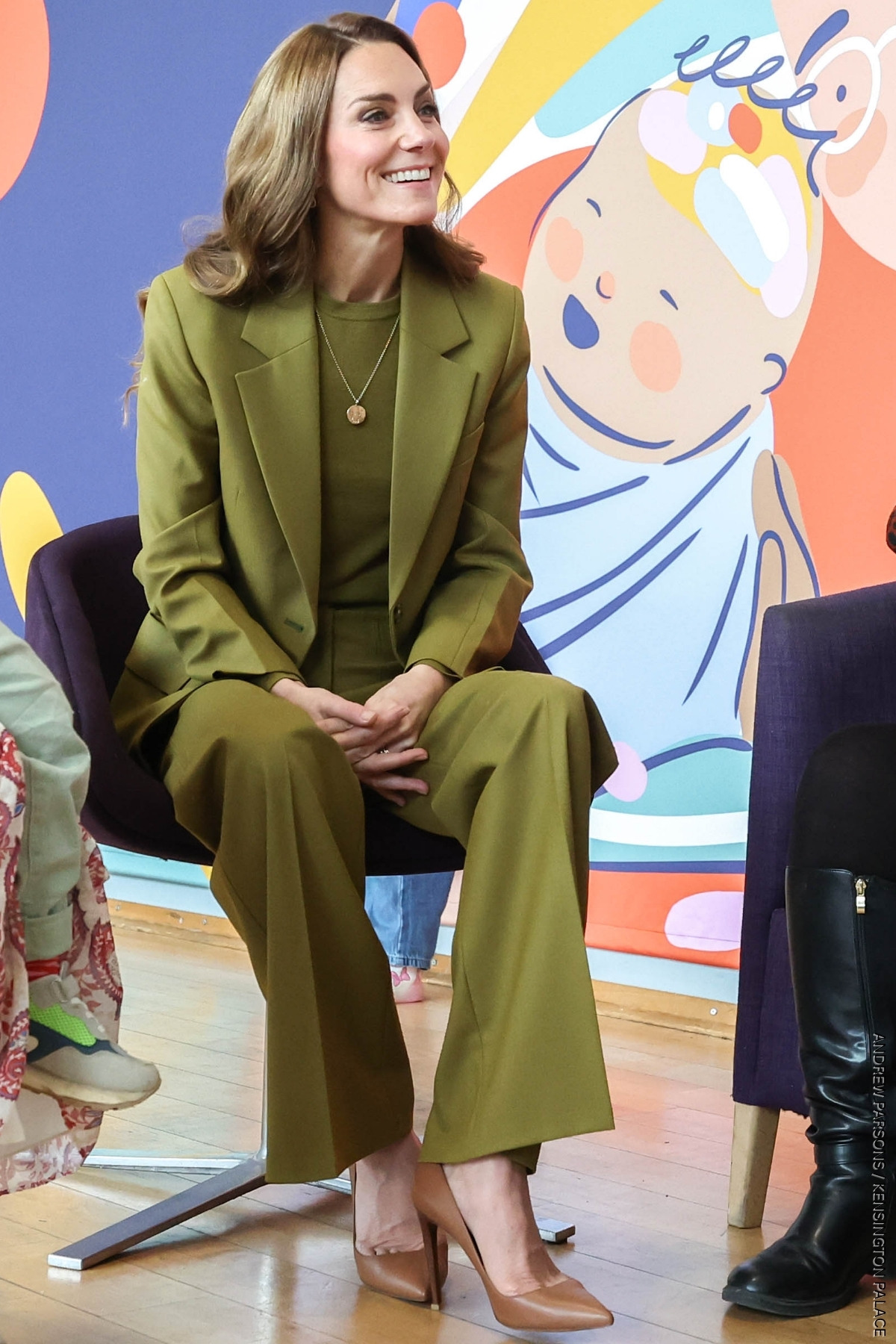 Princess of Wales, seated, wearing an olive green suit with matching olive green sweater.  She finishes the look with a pair of golden brown heels by Ralph Lauren.
