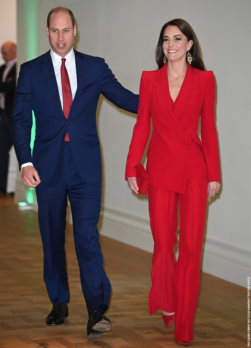 Kate Middleton, smiling and walking with Prince William.  The Princess is wearing a bright red suit by Alexander McQueen. 