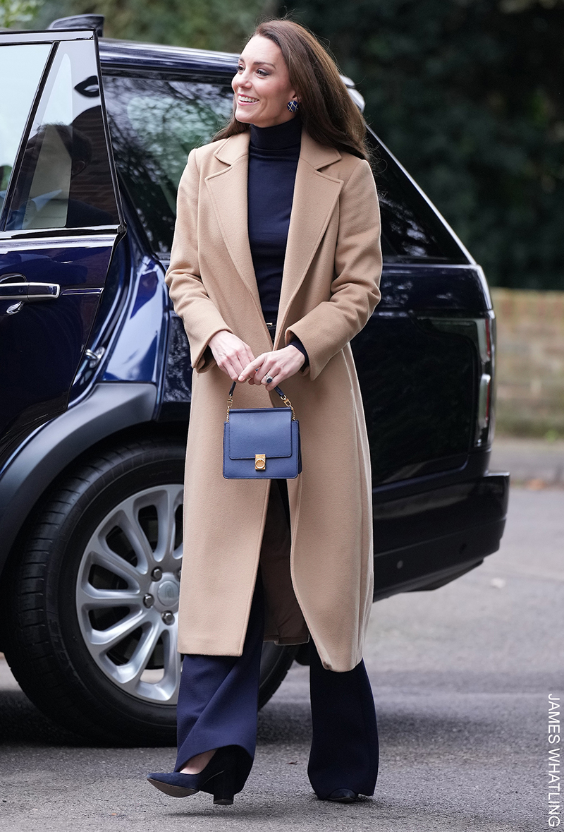 Kate Middleton wearing a pair of navy block heel pumps from Emmy London with blue wide-leg trousers, a long camel coat and a blue handbag