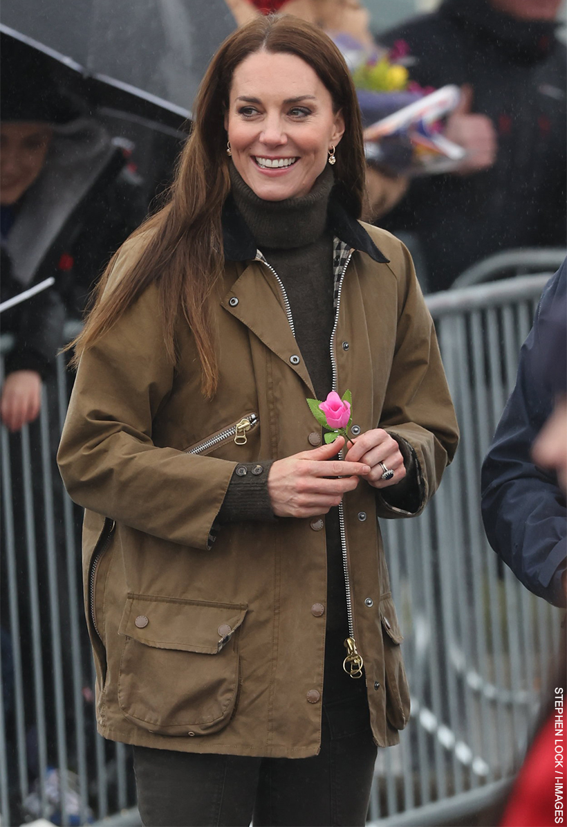 Kate Middleton, the Princess of Wales, stood in her dark olive green Barbour x Alexa Chung Edith jacket, holding a single pink rose. She is smiling.