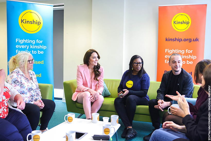 Kate Middleton chatting with people at a Kinship support group.  She's wearing a pink suit with pearl accessories. 