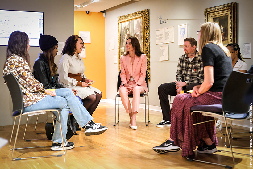 Kate Middleton sitting down with a group of people inside the Foundling Museum.  She's wearing a pink suit with pearl accessories. 