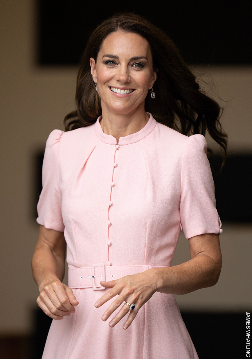 Kate Middleton smiling, wearing a soft pink dress, with her hair curled and blowing in the wind.
