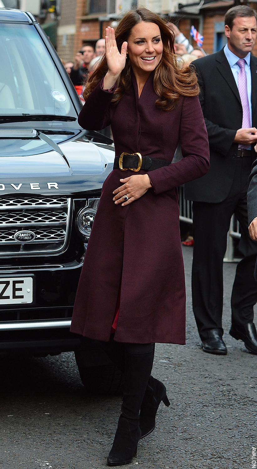 The Princess of Wales wearing a dark aubergine coloured coat, belted at the waist.   Her look is finished with a pair of dark black knee high boots.  She is waving to the public.
