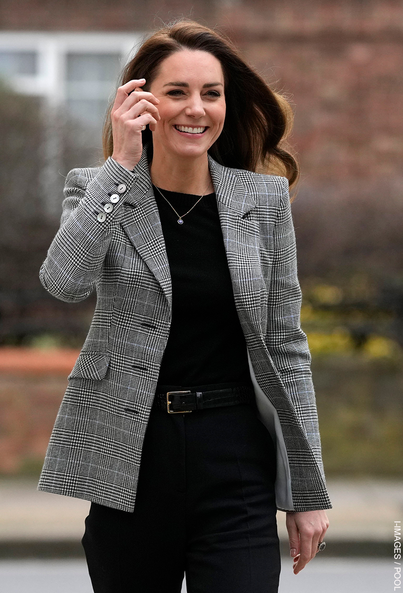 Kate Middleton wearing the glen check blazer over a black sweater.   She is smiling and her hair is blowing in the wind. 