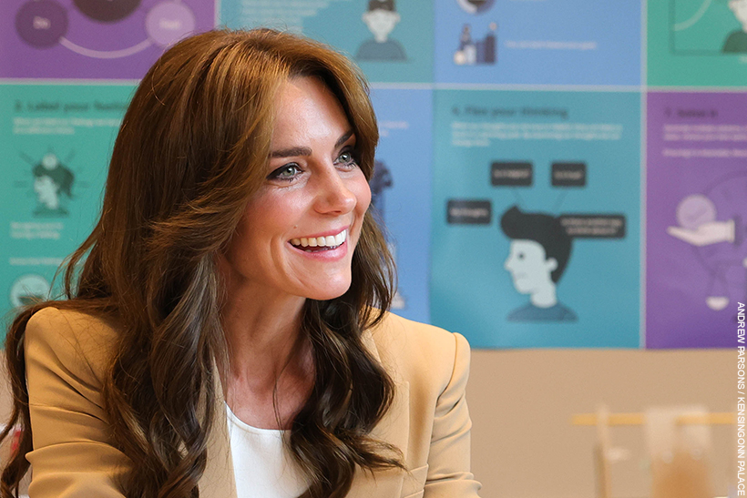 Kate Middleton smiling and engaging in conversation with somebody off camera.  The colourful blue and purple background contrasts with her camel suit and dark curly hair. 