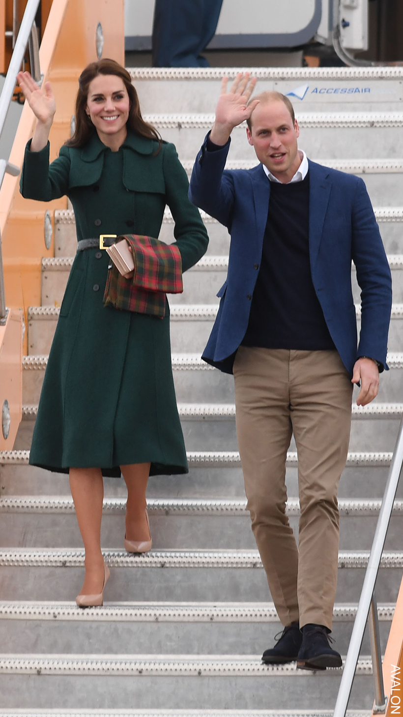 William and Kate disembarking a plane. The Princess is wearing a dark, forest green coat and is holding a green and red tartan scarf. 