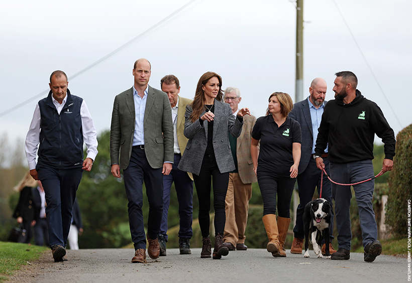 William and Kate stand in the middle of a crowd of people, walking and talking.  Kate is gesturing as she talks.  One of the charity owners, a farmer, walks a black and white collie dog. 