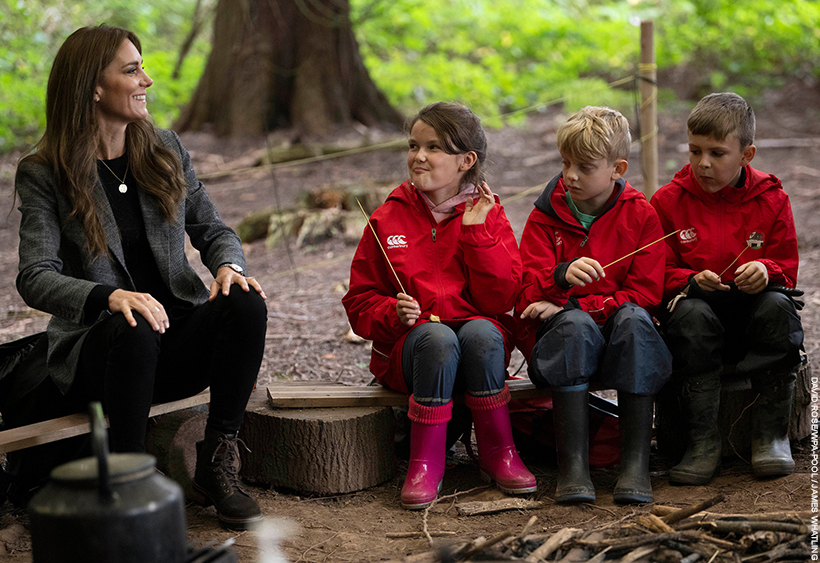 Kate Middleton, dressed in a smart-casual outfit of a grey blazer, jeans and boots, sat on a log chatting with schoolchildren dressed in red.
