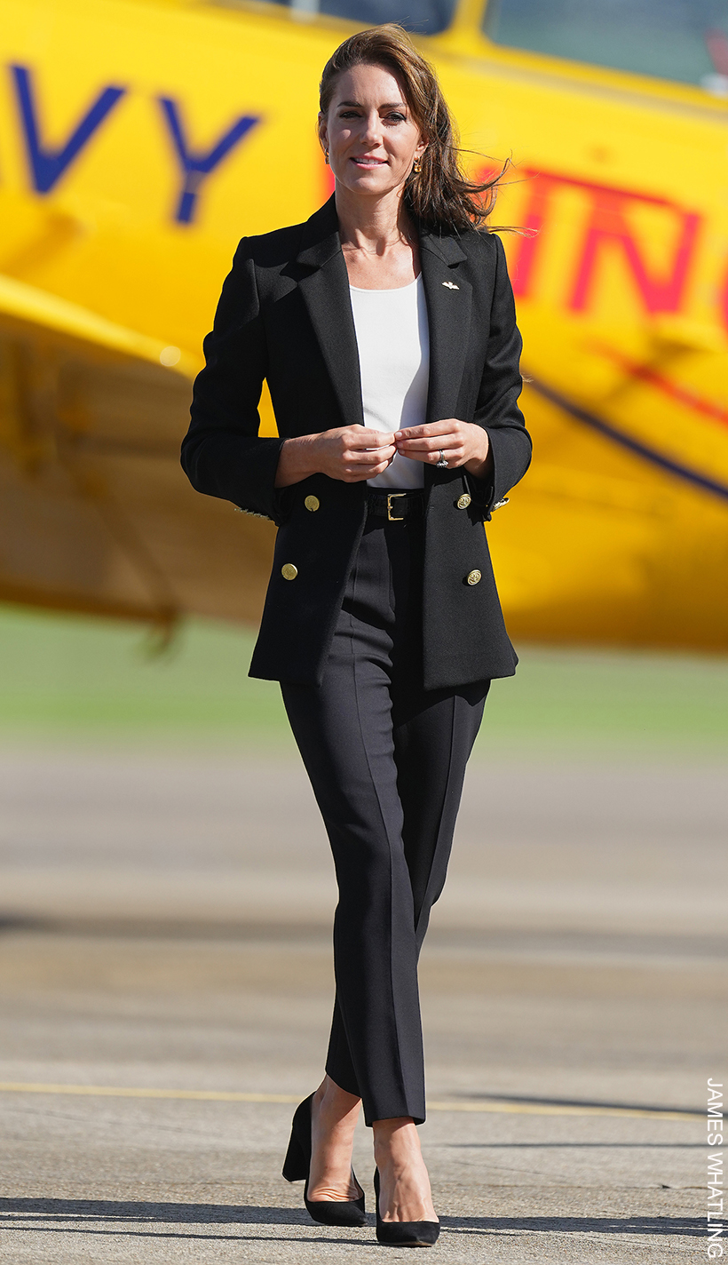 Kate Middleton stood in a black blazer, white top, black trousers and black shoes, in front of of a bright yellow airplane. 