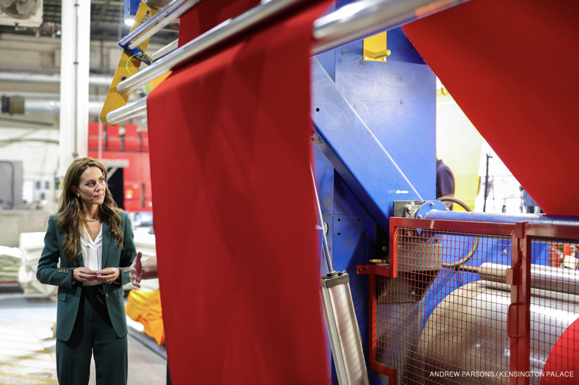 Kate Middleton, wearing a green suit, inspecting a piece of bright red fabric hanging from a textile machine.