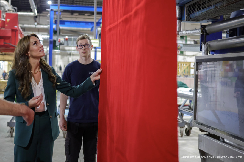 Kate Middleton, wearing a green suit, inspecting a piece of bright red fabric hanging from a textile machine.