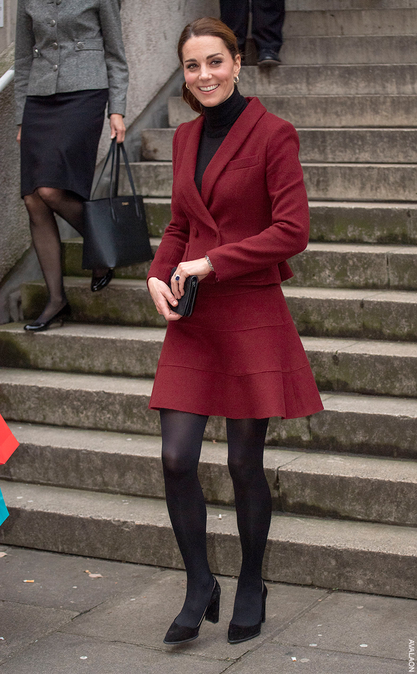 Kate Middleton dressed for the autumn chill in a dark, wine coloured skirt suit over a black turtle neck, with dark tights and dark heels.  Her hair is pulled to the side and she finished her look with gold hoop earrings. 