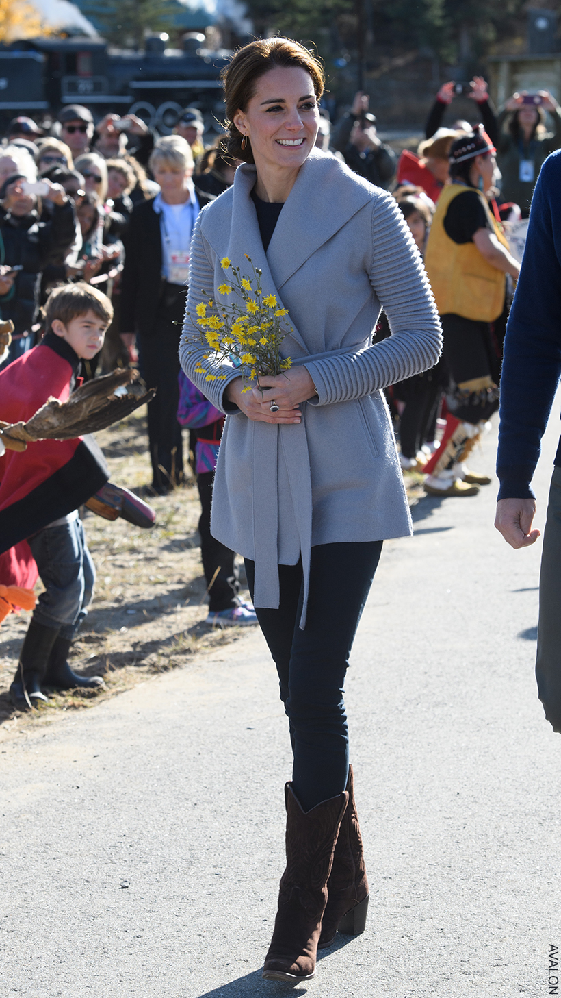 Kate Middleton in a casual grey 'coatigan' over jeans with cowboy boots.  Her hair is up and she's holding a bunch of yellow flowers.