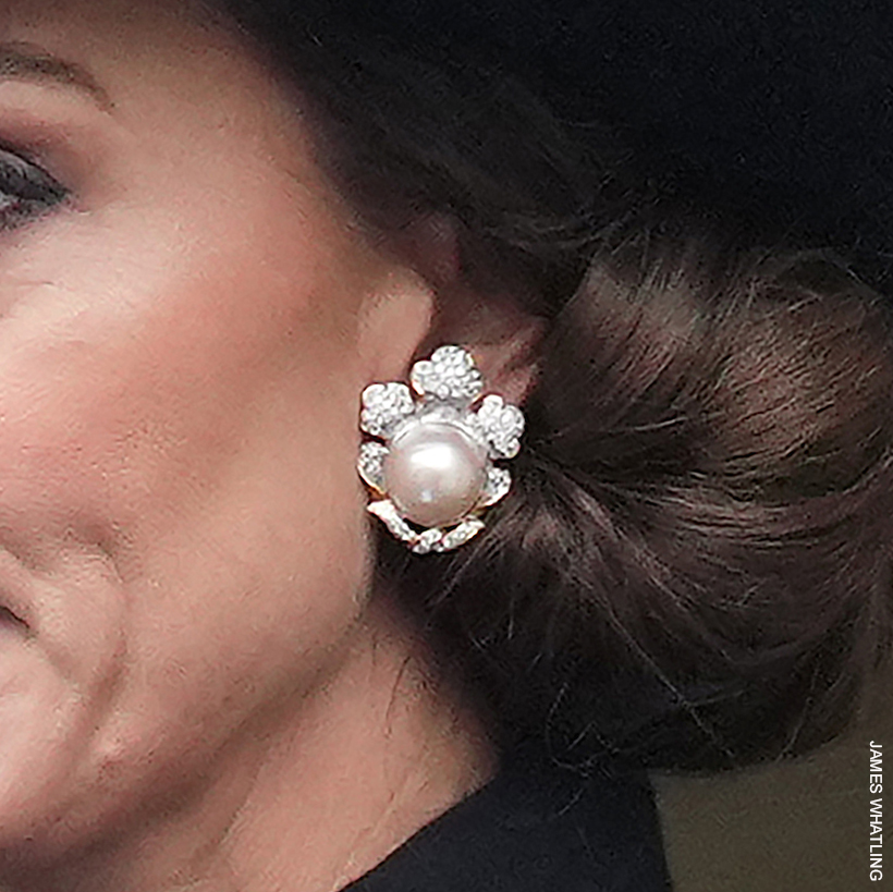 A close-up of a pearl and diamond earrings worn by Kate Middleton, showcasing an intricate floral design with a large central pearl surrounded by diamond petals, set against her low bun hairstyle.