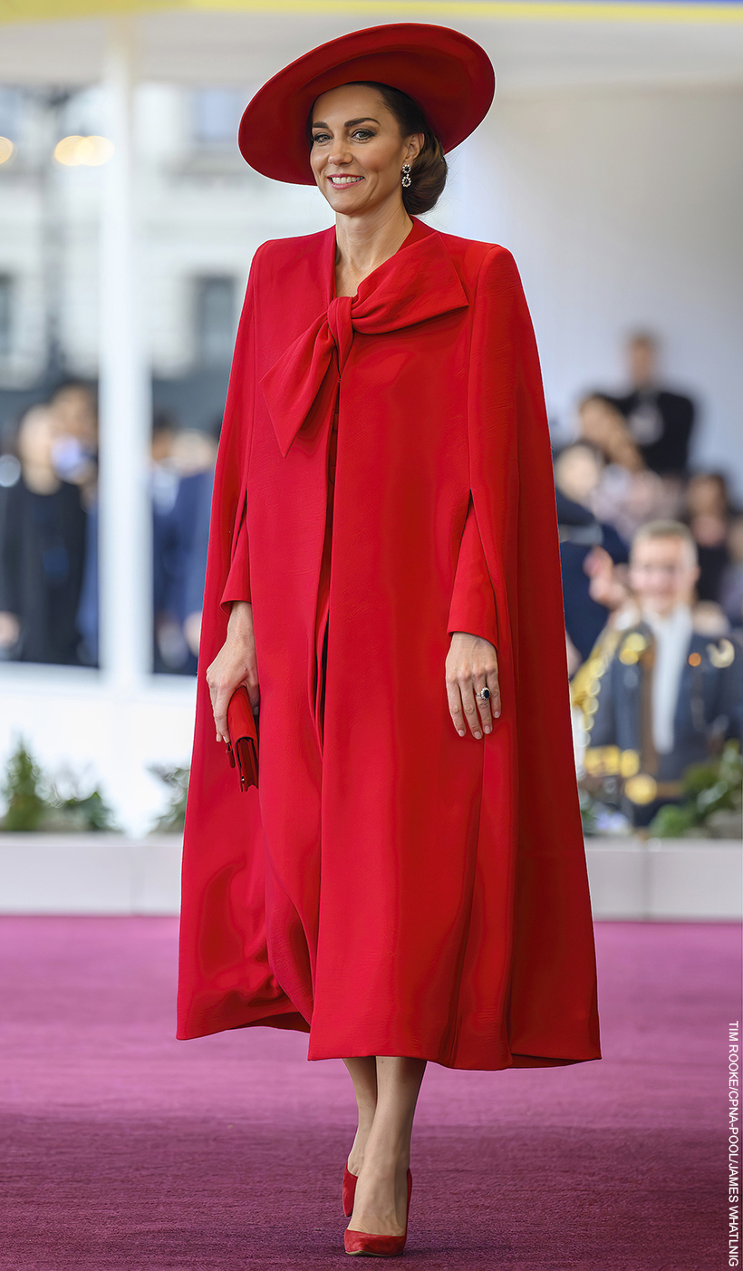 Kate Middleton wears a striking vibrant red cape, featuring a bow, and matching red accessories including a wide brim hat. 