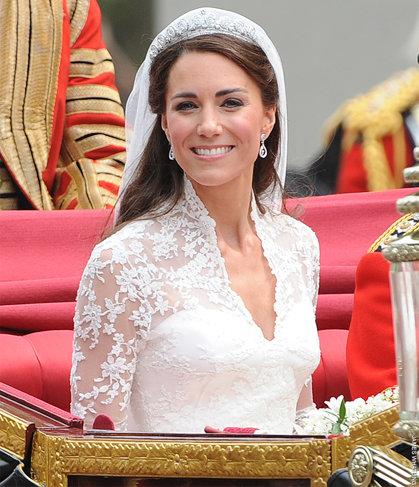 Kate Middleton sat in the carriage on her wedding day, she smiles at the camera, the romantic floral lace on her gown is visible. 