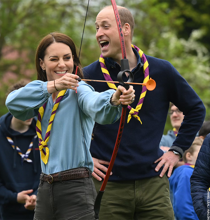The Princess of Wales attempts archery while her husband playfully jeers and laughs behind her. 