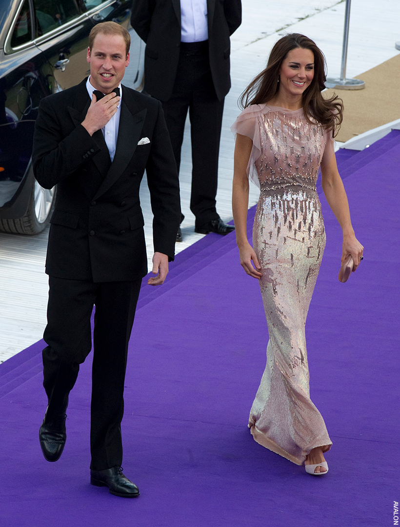 Kate Middleton and Prince William smiling as they walk the purple carpet in formalwear.  The Prince looks dapper in a black tie suit.  Kate wears a shimmering pearlescent pink gown.
