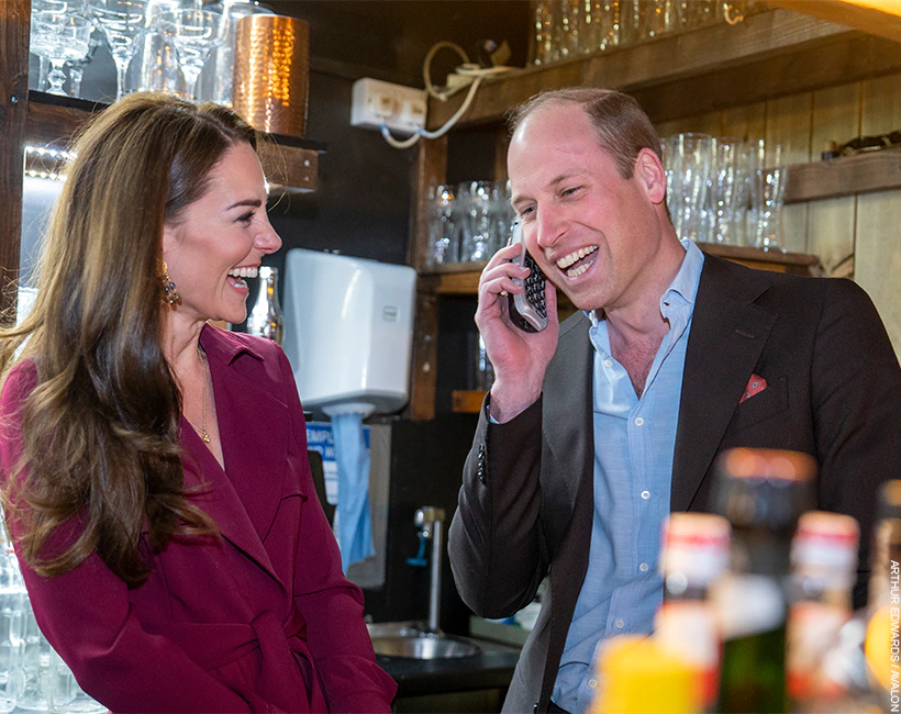 Prince William chats on the phone, as his wife looks on, laughing.  