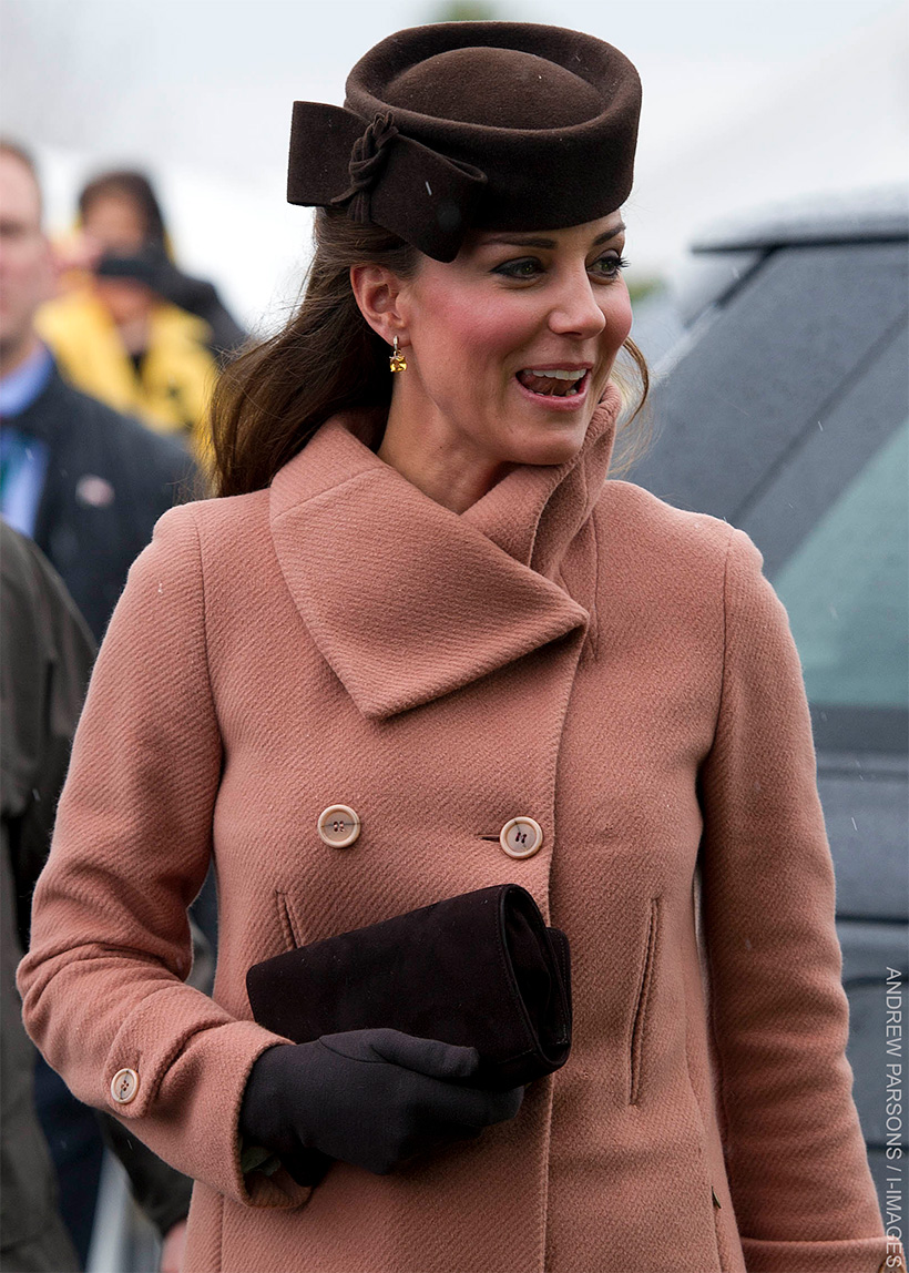 Kate Middleton wearing a salmon-coloured Joseph coat with a wide collar and double-breasted buttons, paired with a brown hat and black gloves, holding a black clutch, during her appearance at the Cheltenham Festival in 2013.