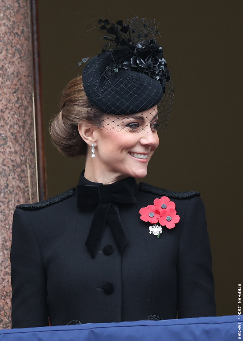 The Princess of Wales stood on the Foreign Office balcony on Remembrance Sunday smiling, wearing a black military-style coat with a bow, a black hat with floral detailing and diamond and pearl earrings