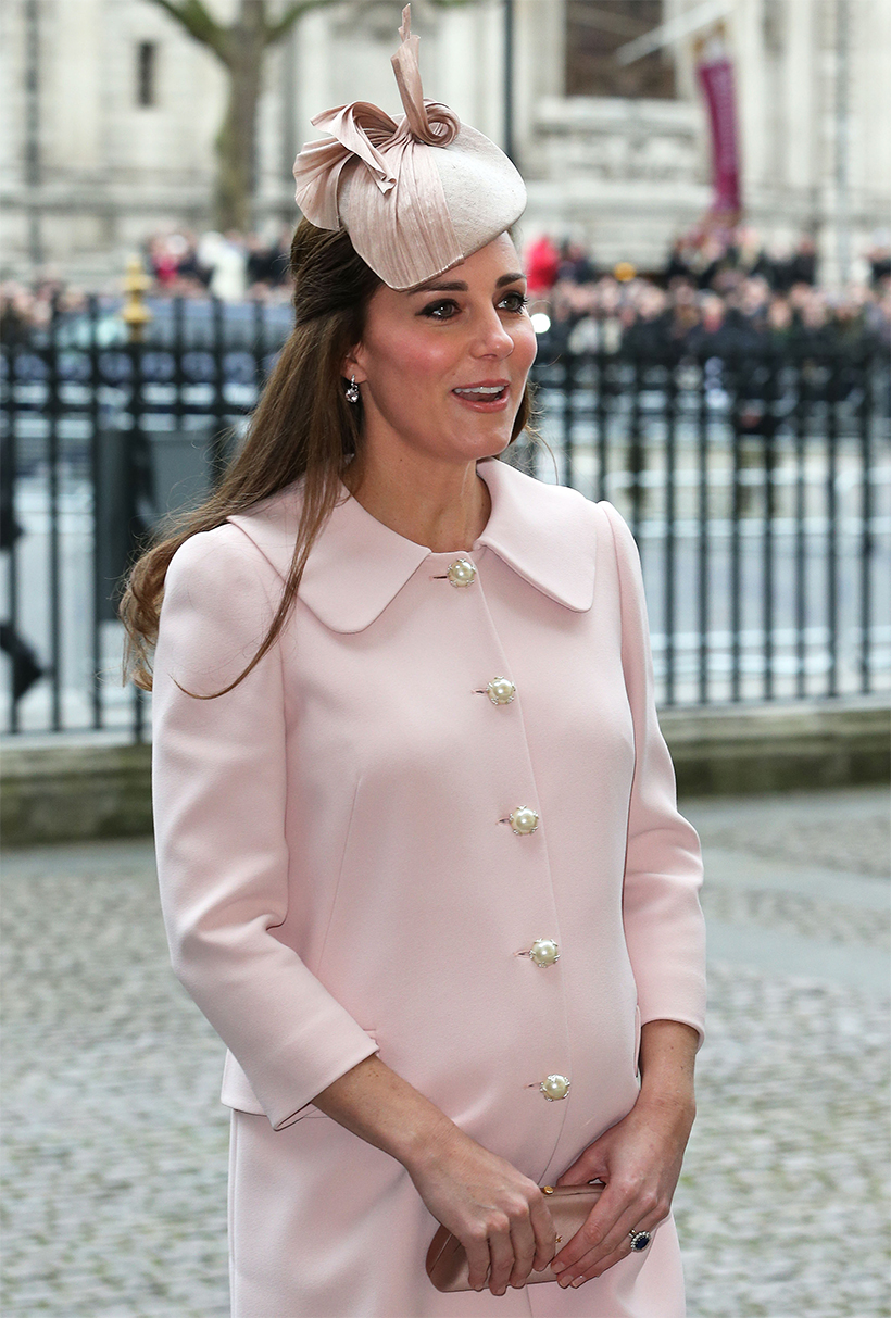 Close-up of Catherine, Princess of Wales (then the Duchess of Cambridge), highlighting her pastel pink coat dress with large pearl buttons, a structured hat with ribbon details, and a nude clutch bag. Her accessories include delicate drop earrings and her signature sapphire and diamond engagement ring. She appears engaged in conversation, with a soft expression on her face. The background is slightly blurred, showing a crowd behind a black iron fence.