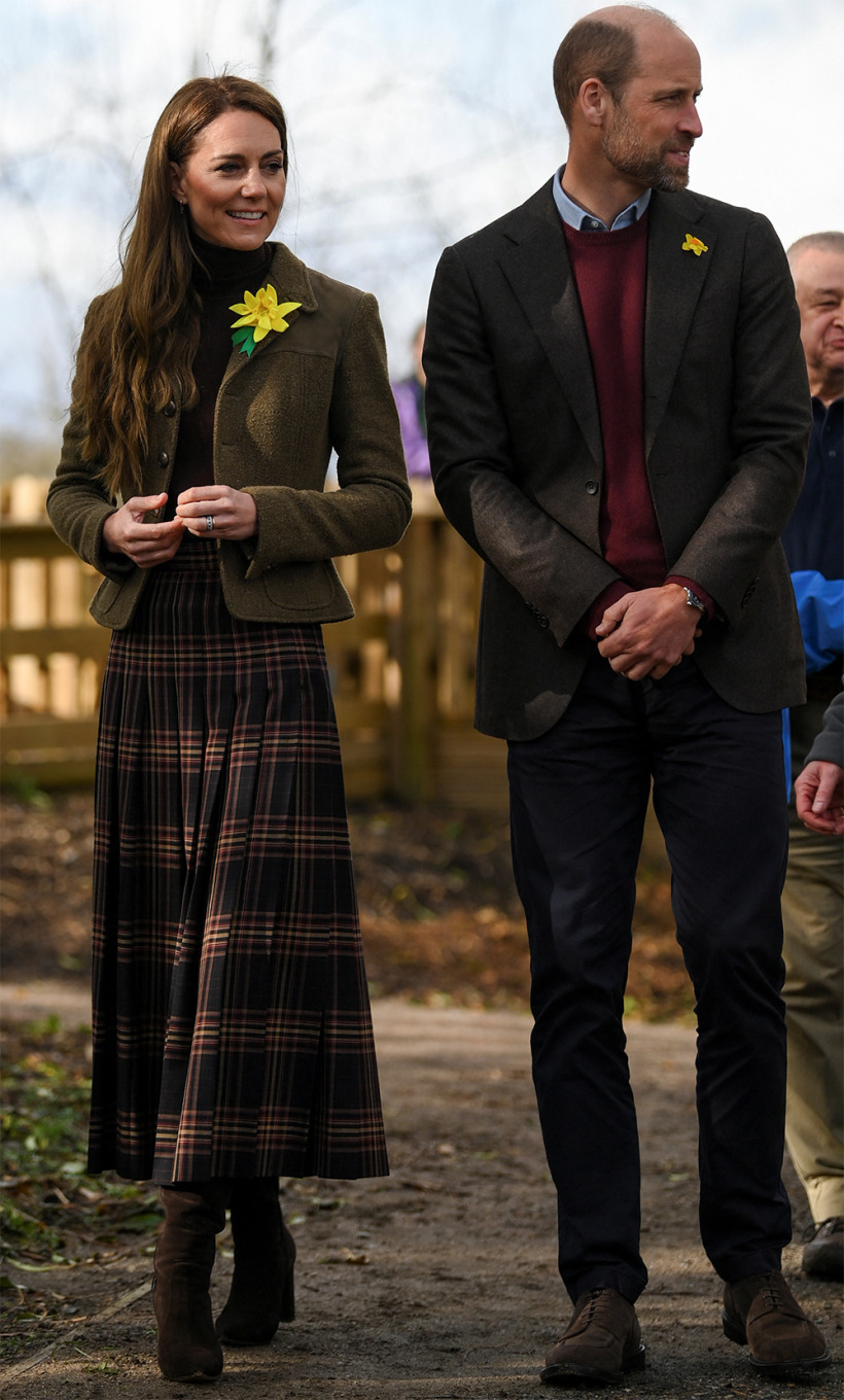 Princess Kate, wearing an olive-green tweed jacket and plaid skirt, smiles warmly while standing next to Prince William, who is dressed in a dark blazer and burgundy sweater. Both are adorned with daffodil pins, symbolizing St. David’s Day, as they engage in conversation at the community garden.