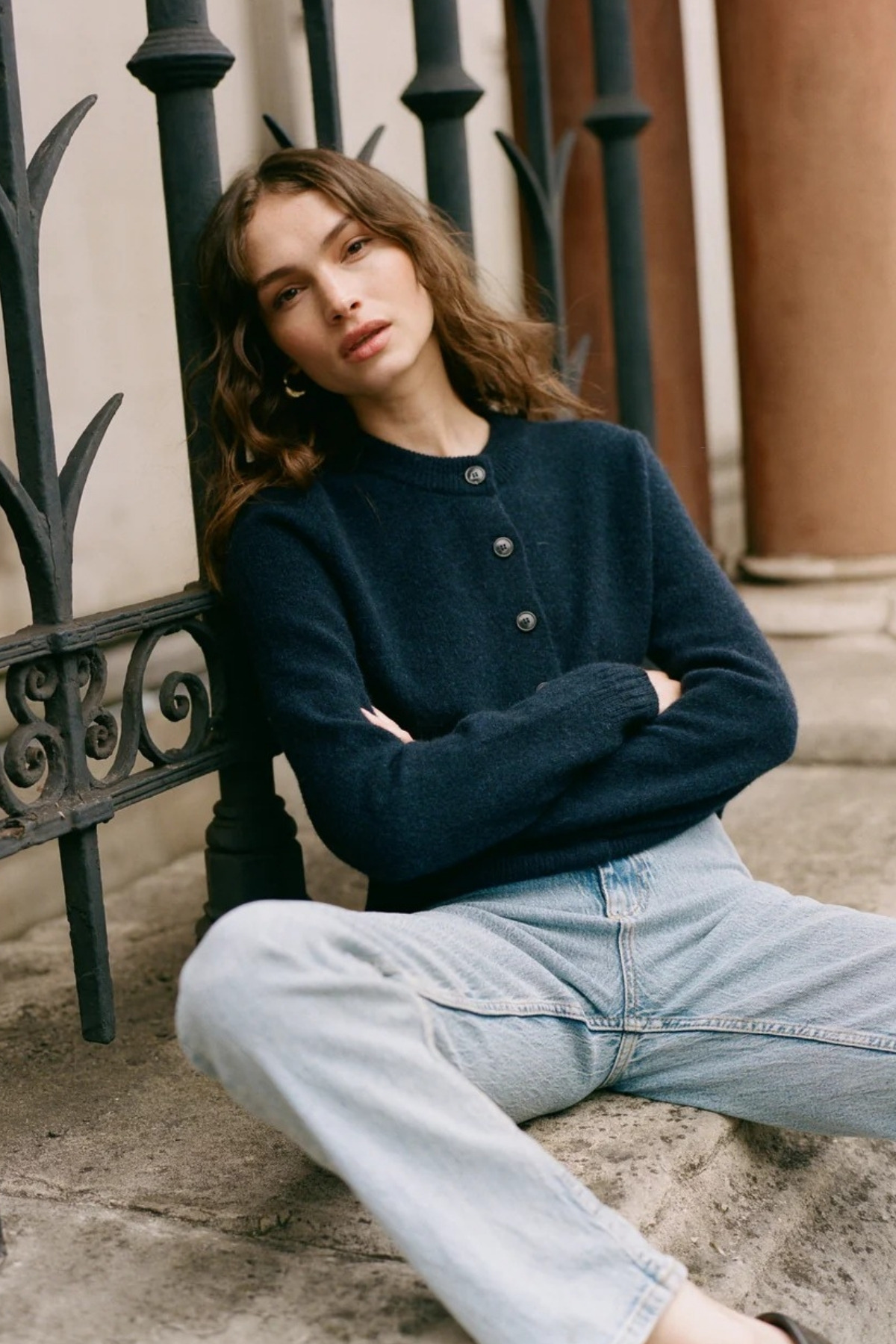 The model is sat down outside, wearing the blue version of the same cardigan, with a light blue pair of jeans.
