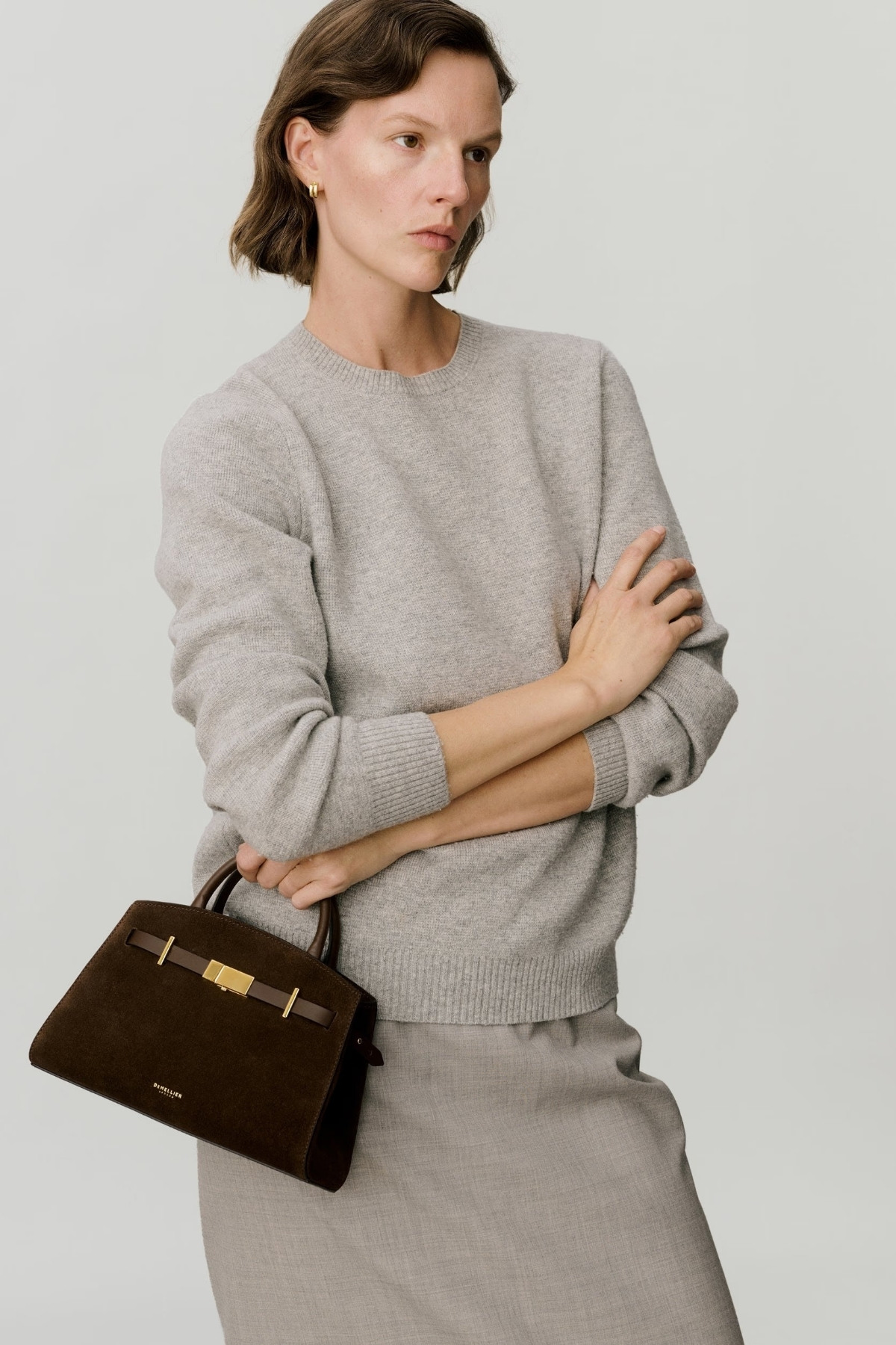 Model with short brown hair wearing grey, carrying the DeMellier Small Hudson in dark brown suede