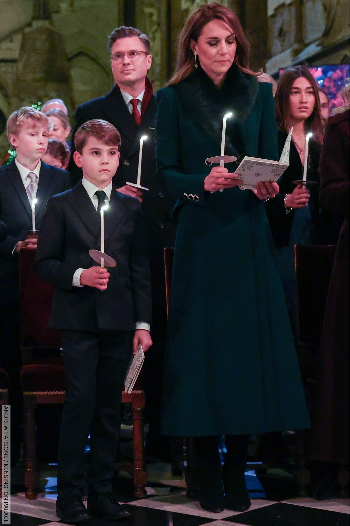 The Princess of Wales, dressed in her green fur topped coat,  at the Christmas Carol service. She is stood with her son Lewis, both holding lit candles.