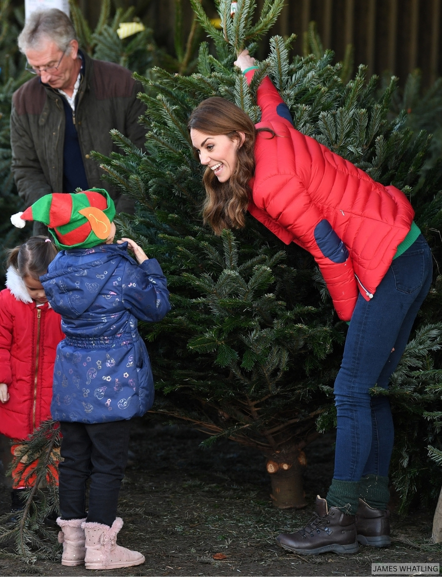 The Princess of Wales, wearing a bold red puffer jacket, reaches for a Christmas tree while talking to a small child